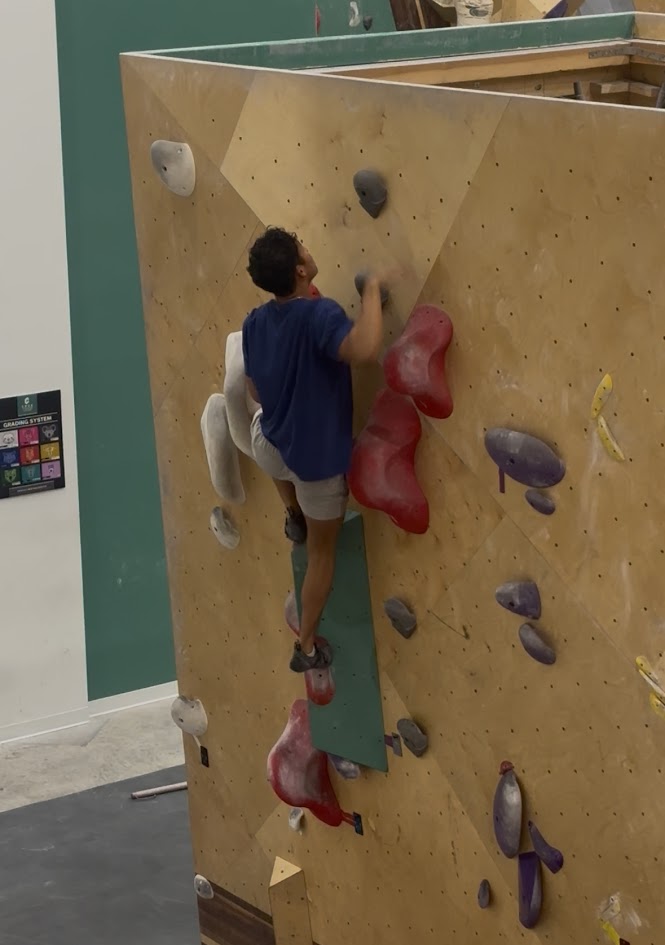 David climbing an indoor bouldering wall with large red and white holds.